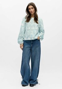 Lichtblauwe blouse met lange mouwen en geborduurd bloemenmotief; gecombineerd met wijde denim jeans met trekkoorden bij de zoom en een ontspannen pasvorm.