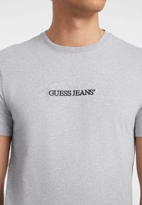 Sivá bavlnená tričko s krátkymi rukávmi a okrúhlym výstrihom, s čiernym vyšitým nápisom "GUESS JEANS" na hrudi.