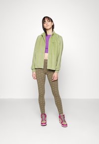 Grøn kordfløjljakke, lilla crop top, olivengrønne leggings med mønster og lyserøde sneakers. Modellen står foran en ensfarvet hvid baggrund.