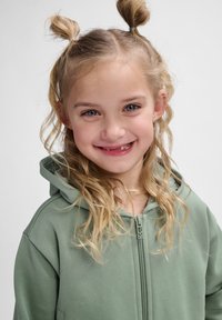 Groene rits-hoodie met een kangoeroe-pocket, zachte stof en een relaxte pasvorm. Model heeft golvend haar en twee knotjes, glimlachend naar de camera.