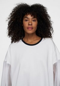 Hvid sweatshirt med lange, gennemsigtige meshærmer og en kontrasterende marineblå ribkant. Enkel design, blød tekstur og et minimalistisk udseende.