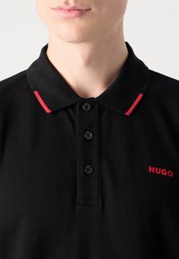 Čierna polokošeľa s textúrovanou látkou, trojiciou gombíkov a červeným pruhom na golieri, s červeným logom "HUGO".
