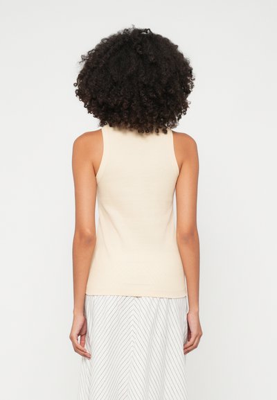 Svetlo bež brezrokavni top iz mehkega, rebrastega materiala. Prilegajoča oblika z okroglim izrezom, izpostavljenimi šivi in gladko teksturo.