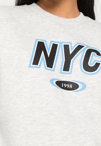 Sudadera gris con "NYC" en letras negritas de color negro y azul claro, con "1998" en negro rodeado por un óvalo azul. Textura de tela suave.