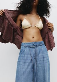 Lys gul bikini-top med bølgede kanter, parret med høj talje blå denim bukser. En løs, burgundy cover-up holdes åbent.