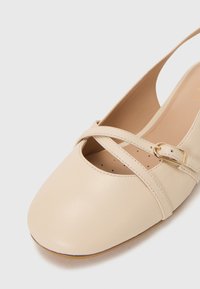 Beige lädersko med rund tå och utskårna detaljer. Har korsade remmar med en liten guldbuckle och en slät yta.