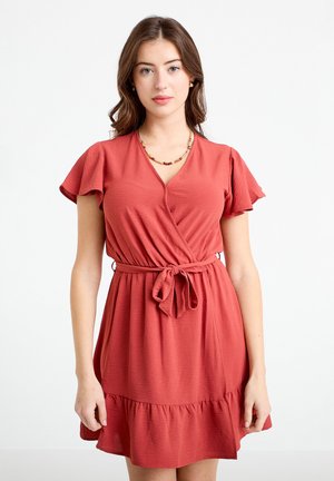 Ung kvinde med lys hud og brunt bølget hår iført en rustfarvet slå-om-kjole med korte ærmer, bundet i taljen, og med en perledekoration halskæde.