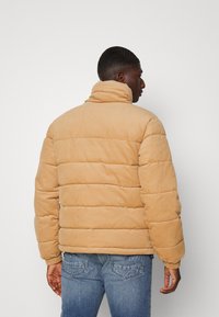 Beige cordfløyel pufferjakke med høy krage, quiltet design og ribbede mansjetter. Brukt over blå jeans, med en avslappet passform og tekstur.