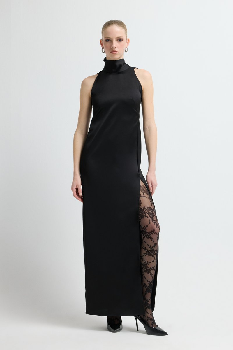 Czarna satynowa sukienka na szyję z wysokim rozcięciem, odsłaniająca przezroczyste czarne koronkowe rajstopy, zestawiona z czarnymi szpilkami. Gładka faktura, dopasowany fason.
