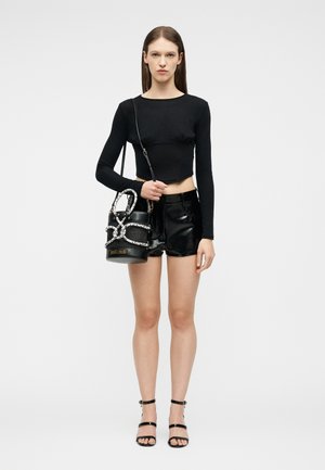 Femme portant un crop top noir à manches longues, un short noir brillant, des talons noirs à bretelles, tenant un sac à main noir avec des chaînes argentées.