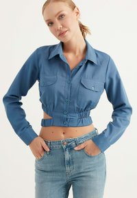 Camicia blu cropped realizzata in tessuto morbido, con maniche lunghe, chiusura frontale con bottoni e due tasche sul petto con bottoni abbinati.