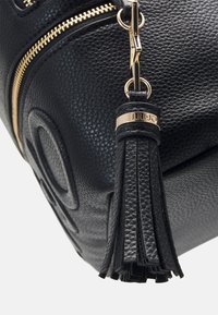 Schwarze Handtasche mit strukturiertem Material, goldener Reißverschluss, Lederquaste und Logodetail am Henkel. Nahaufnahme der Seitenansicht.