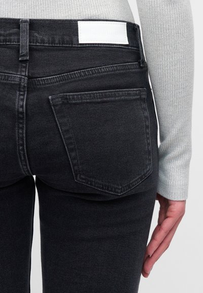 Κοντινή λήψη της κάτω πλάτης ενός ατόμου που φοράει μαύρο τζιν με πίσω τσέπη και λευκή ετικέτα, μαζί με ένα ανοιχτό γκρι πλεκτό μακρυμάνικο μπλουζάκι.