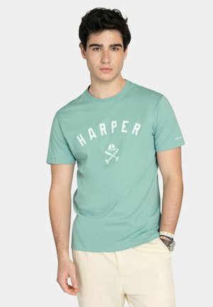 Joven vestido con una camiseta de color verde agua clara con la palabra "HARPER" y dos bates de cricket cruzados, y pantalones crema, de pie con una mano en el bolsillo.