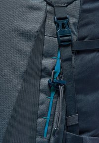 Zaino grigio scuro in tessuto con sezioni testurizzate, dettagli in teal e chiusura a fibbia. Spallacci regolabili con cordino elastico e fermaglio.