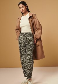 Hnedá kapucňová bunda cez krémový top, v kombinácii so zvieracími nohavicami s leopardím vzorom. Model má na sebe svetlé tenisky. Pozadie je jednoducho pieskovej farby.