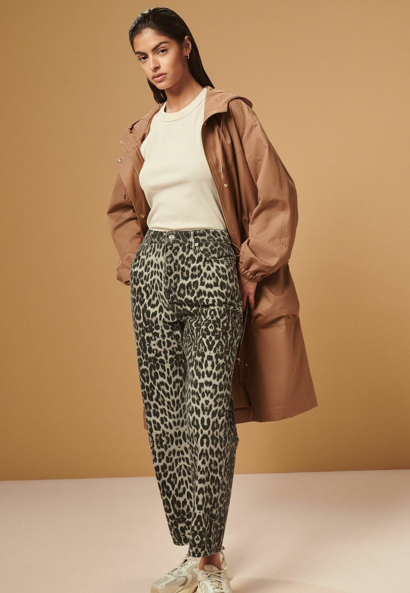 Giacca cappuccio marrone sopra una maglietta crema, abbinata a pantaloni leopardati. La modella indossa sneaker chiare. Lo sfondo è di un beige uniforme.