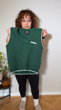 Grønn strikket vest med V-hals, hvit kant langs kantene og en logo på fronten. Ribbet kant nederst.