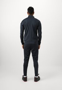 Tumesin sinine spordisügis, millel on lukuga jakk ja kitsenevad püksid, valmistatud siledast ja venivast kangast, koos spordijalanõudega.