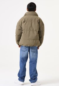 Chaqueta de plumas en verde oliva, con corte oversized, caracterizada por un cuello alto y un diseño acolchado. Combinada con jeans azules holgados y zapatillas blancas.