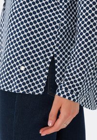 Blusa bianca con pois blu marine, caratterizzata da un orlo curvo, fessure laterali e un singolo bottone bianco in basso.