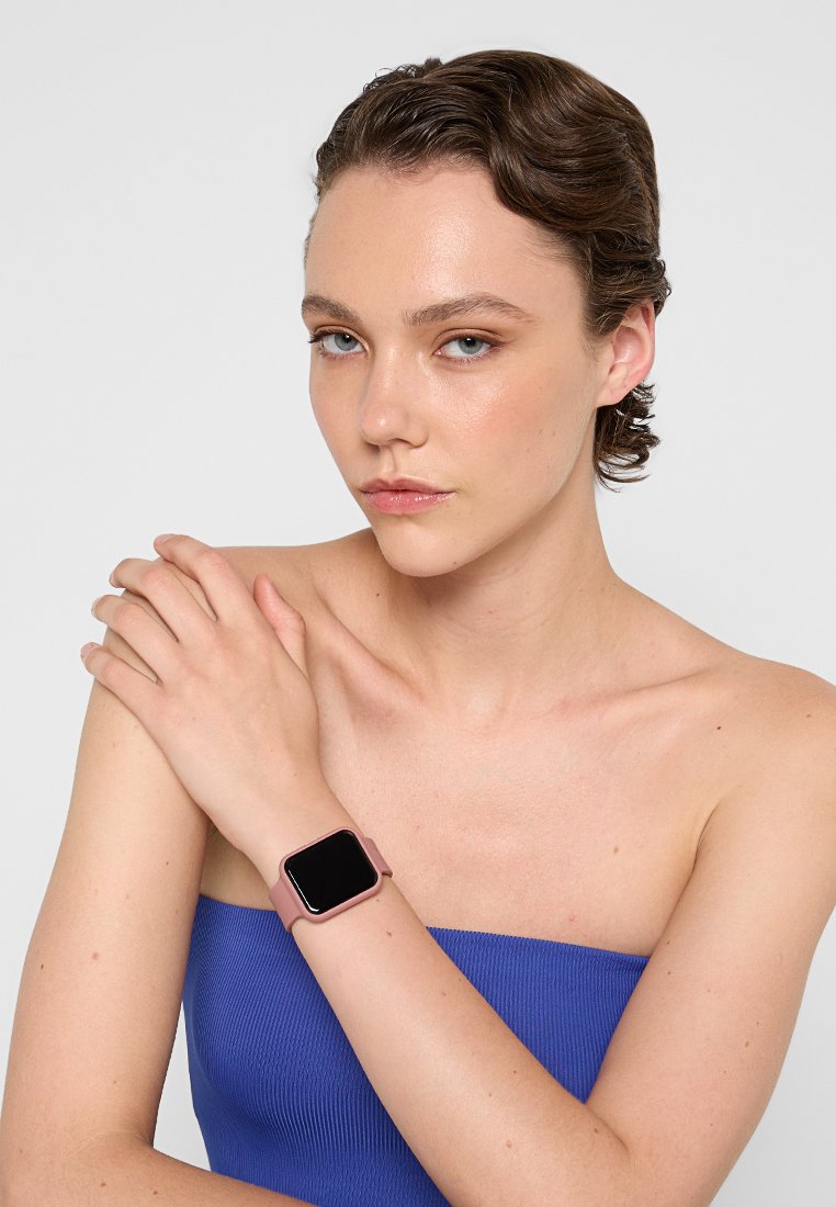 Montre connectée avec un cadran carré, affichage noir et bracelet en silicone rose clair. Le modèle porte un haut côtelé bleu, affichant la montre sur son poignet.