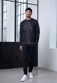 Chaqueta bomber de cuero negro sobre un suéter de punto verde oscuro, combinada con pantalones a rayas y zapatillas blancas. Estilo moderno y minimalista.