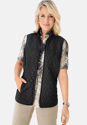 Gilet trapuntato nero con chiusura a zip e tasche laterali, indossato sopra una camicia a maniche corte a fantasia. Presenta un tessuto testurizzato e colletto alto.