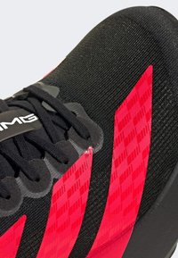 Scarpa sportiva nera con accenti geometrici rossi, tomaia testurizzata, lacci a contrasto e una suola in gomma liscia. Mette in risalto i dettagli del design sportivo.