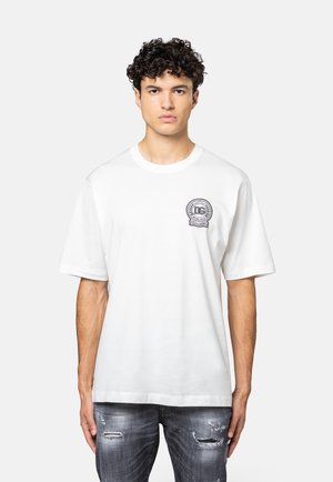 Jonge man met krullend haar die een wit T-shirt met korte mouwen draagt met een zwart DG-logo op de borst en versleten grijze spijkerbroek, rechtop staand.