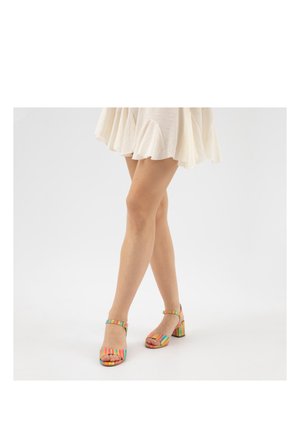 Frau trägt Sandalen mit Absatz in Pastell-Regenbogenstreifen und Knöchelriemen sowie einen kurzen, cremefarbenen Rock.