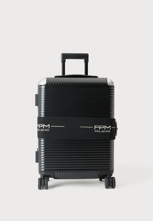 Valise rigide noire avec quatre roues et une poignée étendue, sécurisée par une sangle noire portant la mention FPM Milano.