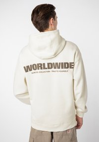 Cremefarbener Hoodie mit einem großen "WORLDWIDE"-Grafik in Braun auf der Rückseite. Mit einer Kapuze mit Kordelzug und gerippten Bündchen. Lässiger Fit.