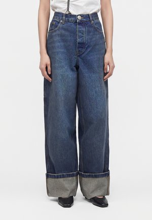 Person iført vidde blå jeans med store opslag, kombineret med sorte sko med firkantet tå og en hvid, indstukket top.
