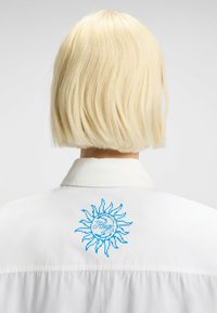 Witte shirt met een blauw zonnestraalontwerp met de tekst "Hugo" gecentreerd binnen de cirkel, gemaakt van soepel materiaal met een kraag.
