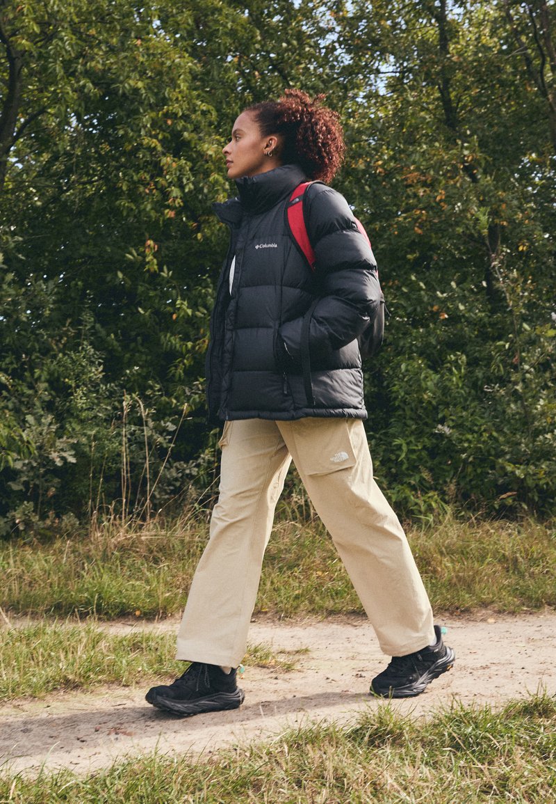 Czarna kurtka puchowa, beżowe szerokie spodnie, czarne obuwie. Plener z zielenią, swobodnie spacerując wzdłuż ziemistej ścieżki.