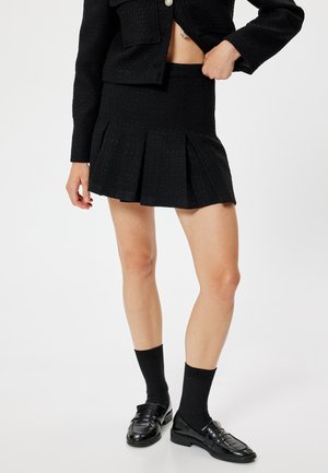 Zwarte geplooide minirok met een gestructureerd patroon; aangesloten bij de taille, gedragen met zwarte sokken en schoenen, gepresenteerd tegen een lichte achtergrond.