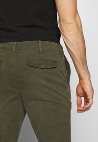 Olive green cotton chinos with a buttoned back pocket, visible stitching details, and a straight leg cut. Fitted waist and smooth texture.