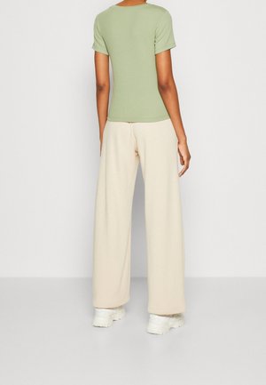 Personne debout, de dos, portant un haut vert clair à manches courtes, un pantalon beige à jambes larges, et des baskets blanches sur un fond uni.