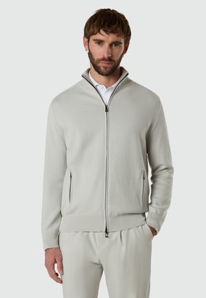 Männliches Model mit kurzen braunen Haaren und Bart, trägt eine hellgraue Jacke mit Reißverschluss über einem weißen Hemd mit Kragen sowie passende hellgraue Hosen.