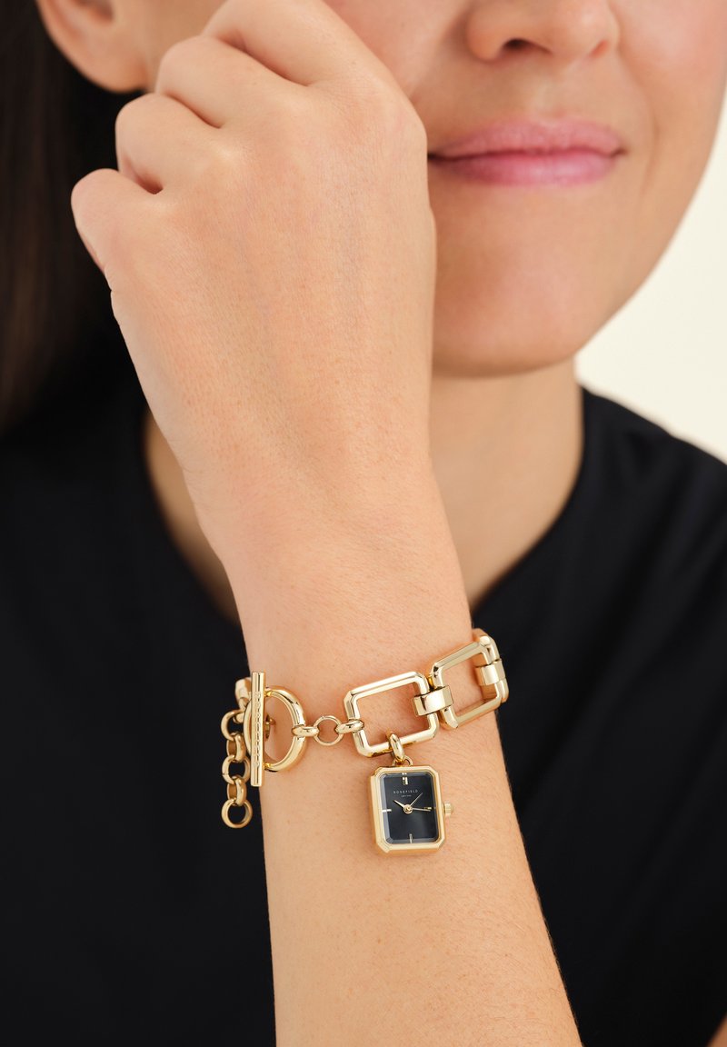 Reloj de pulsera en tono dorado con caja rectangular, esfera oscura, diseño de cadena unida y cierre de palanca. Textura suave y acabado pulido.