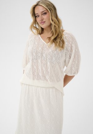 Woman with wavy blonde hair wearing a white, patterned, knitted top and matching skirt, standing against a plain light background.
