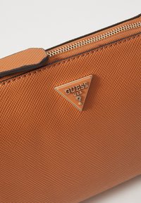 Sac à main en synthétique texturé orange avec fermeture éclair dorée et logo en métal triangulaire arborant "GUESS USA." Accentué de garnitures brunes.