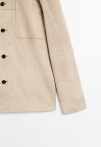 Beige suede jacket with a collar, button closure, front pocket, and long sleeves. Smooth texture and tailored fit.