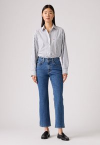 Prúžkovaná košeľa s button-up v bielej a námorníckej modrej farbe, kombinovaná s modrými rozšírenými džínami. Čierne mokasíny dopĺňajú outfit. Jednoduchý, neformálny dizajn.