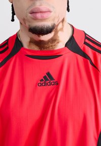 Red adidas sports jersey with black accents, textured vertical lines, and a prominent black logo on the front. Short-sleeved design.
