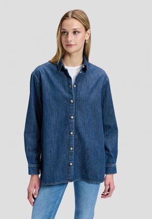 Kvinde iført en løs mørkeblå denimskjorte med knapper over en hvid top og lyseblå jeans mod en lys baggrund.