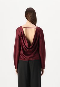 Burgundy satin top with long sleeves, scoop back, and draped detail, featuring a thin strap across the upper back.