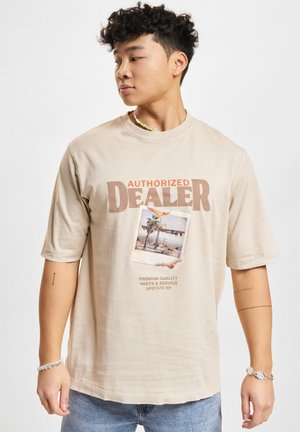 Beige T-Shirt mit kurzen Ärmeln, auf dem der Text "AUTORISIERTER HÄNDLER" in Braun darauf steht, sowie ein Fotodruck von Palmen und einem Text darunter.