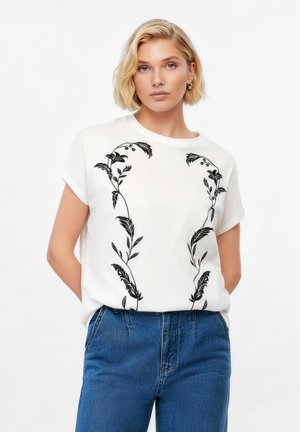 Blonde vrouw die een wit shirt met korte mouwen en zwarte bloemenborduur draagt, gecombineerd met een hoge taille blauwe spijkerbroek, staand met haar handen achter haar rug.
